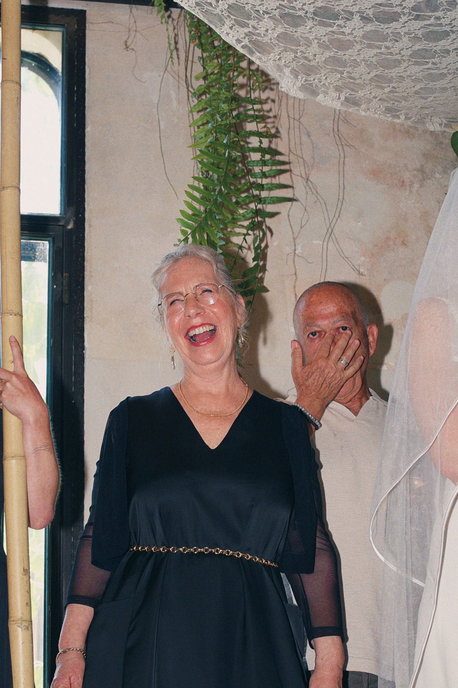 Elderly woman in black dress laughs joyfully at wedding ceremony under chuppah.