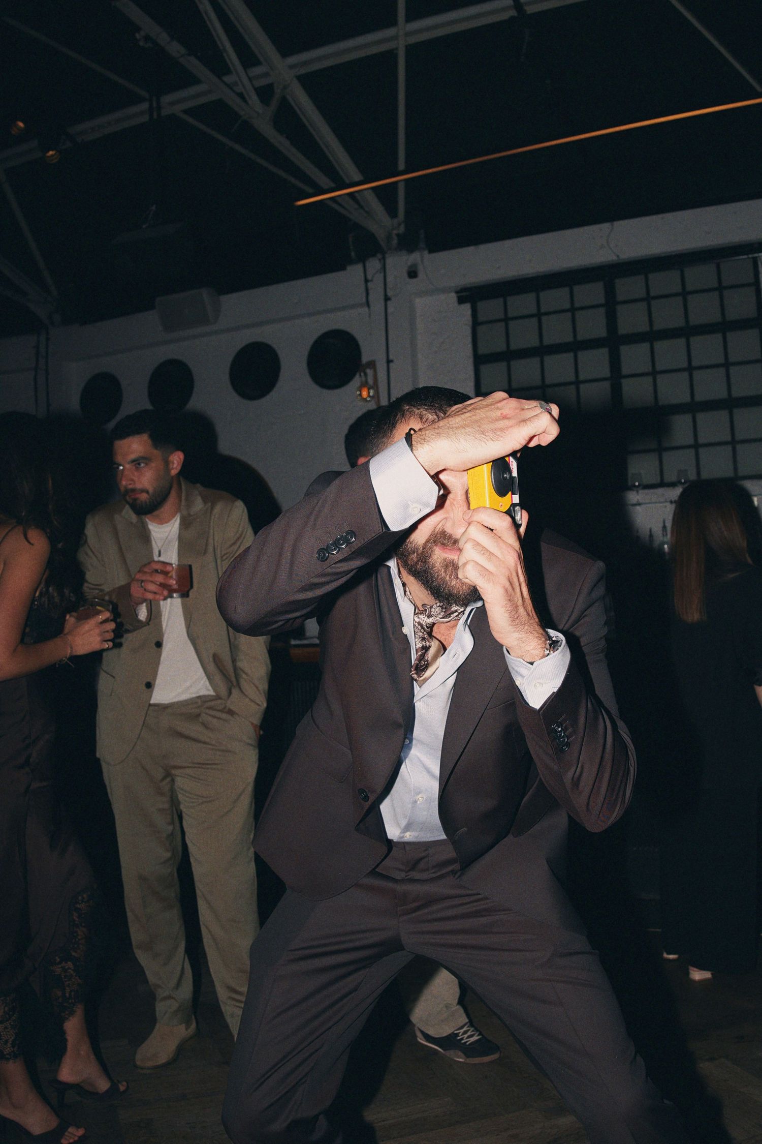 Guest in brown suit photographs with yellow camera while dancing at dark indoor wedding reception.