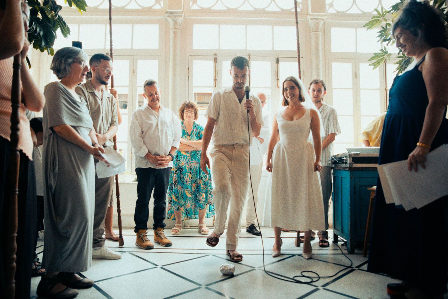 Wedding guests gather in a bright sunlit room with white decor and large windows for a celebration.