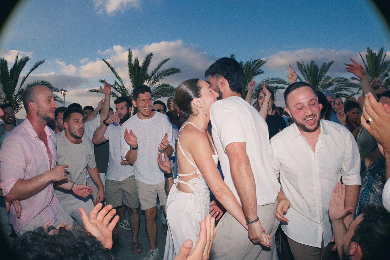 Bride and groom share a kiss surrounded by cheering guests at outdoor wedding party.