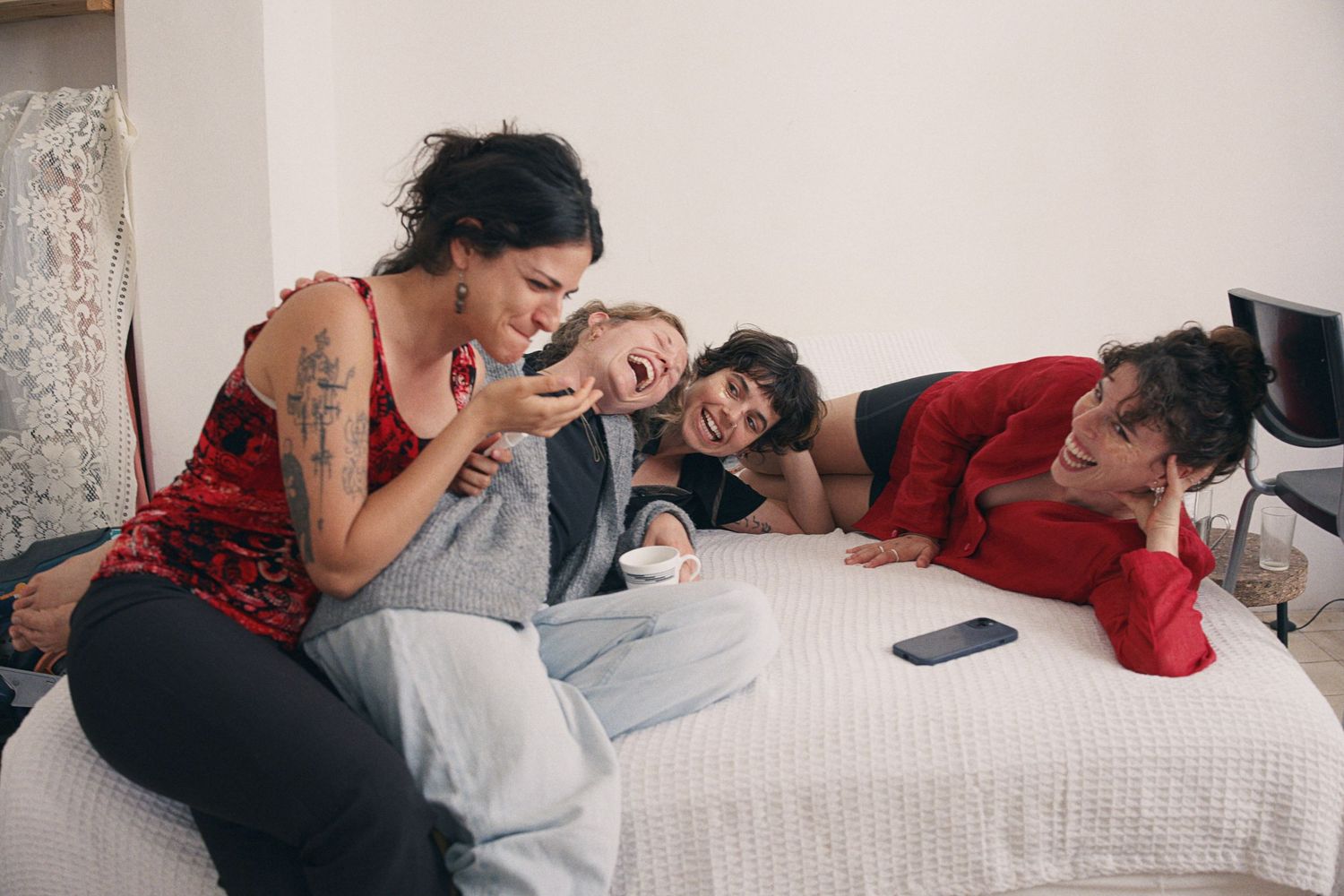 Four friends laughing and having fun together on a bed, sharing a joyful moment while looking at a smartphone.