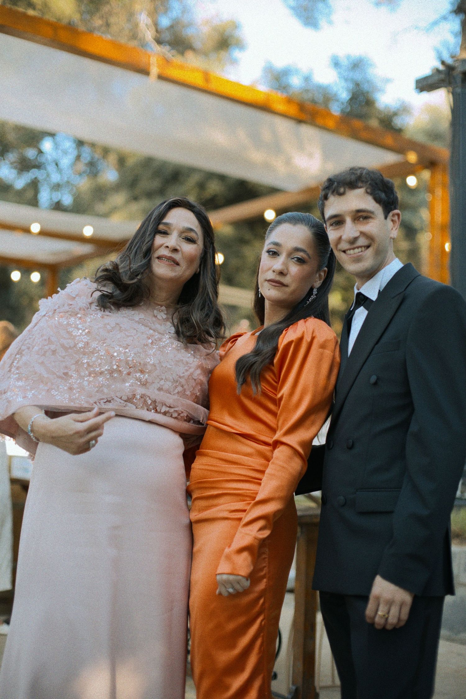 Wedding party members in formal attire including black suits and orange and pink dresses pose together outdoors.
