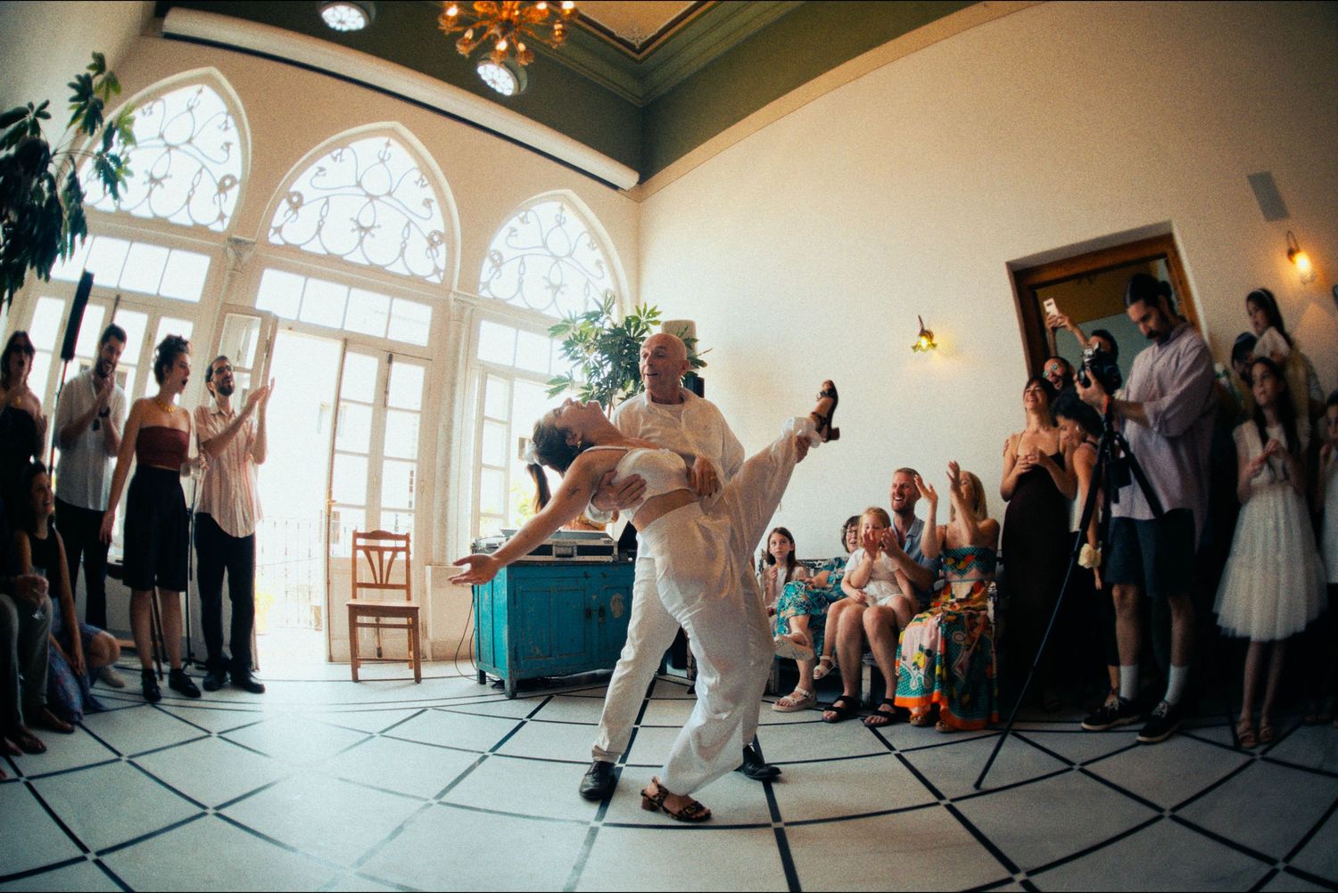 Dancers perform on a checkered floor during a wedding reception in an elegant room with arched windows.