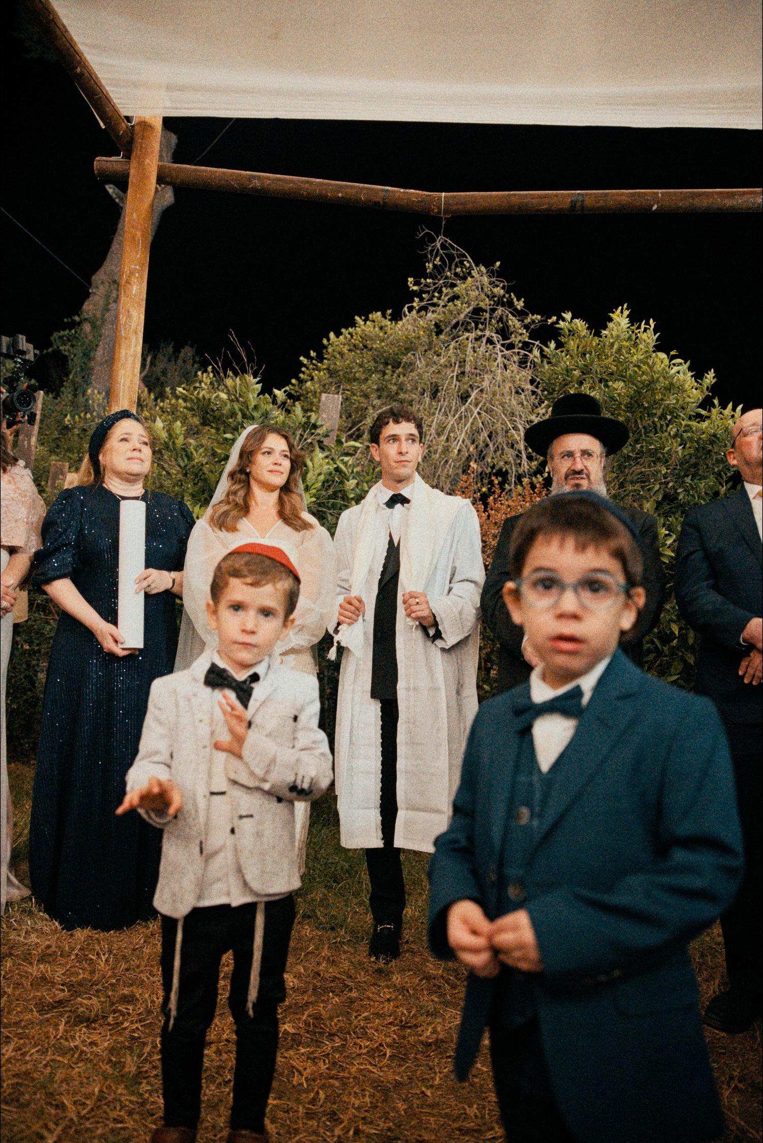 Young attendants in formal attire gather at an outdoor wedding celebration under string lights.