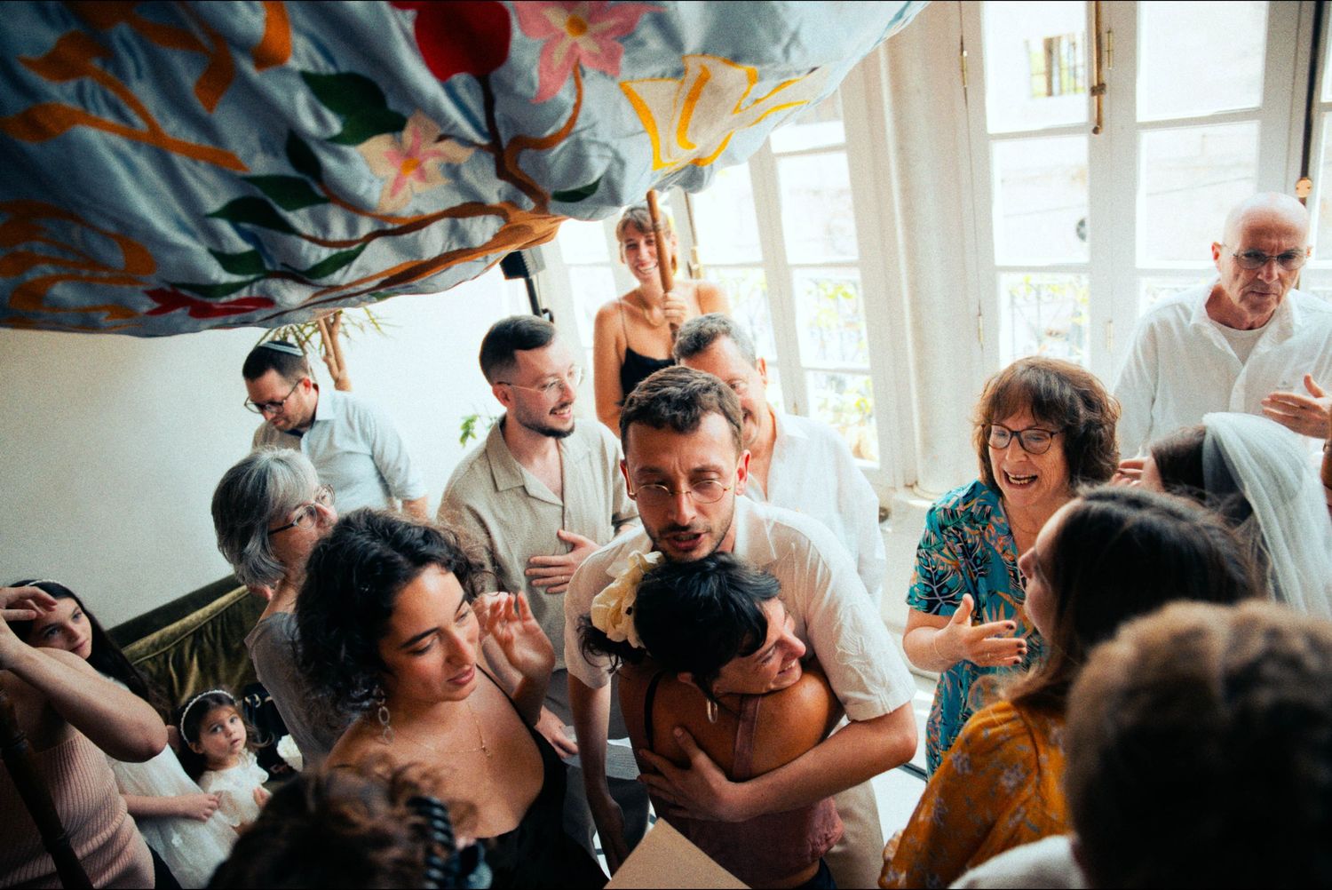 Wedding guests laugh and celebrate under colorful ceiling decorations at an indoor reception.
