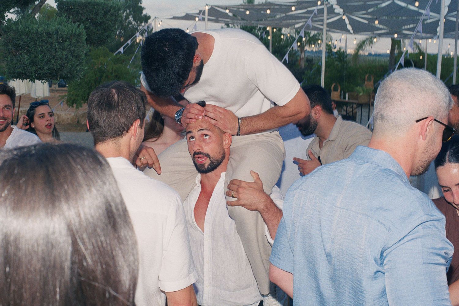 Guest playfully blesses man at crowded outdoor wedding party with palm trees visible in background.
