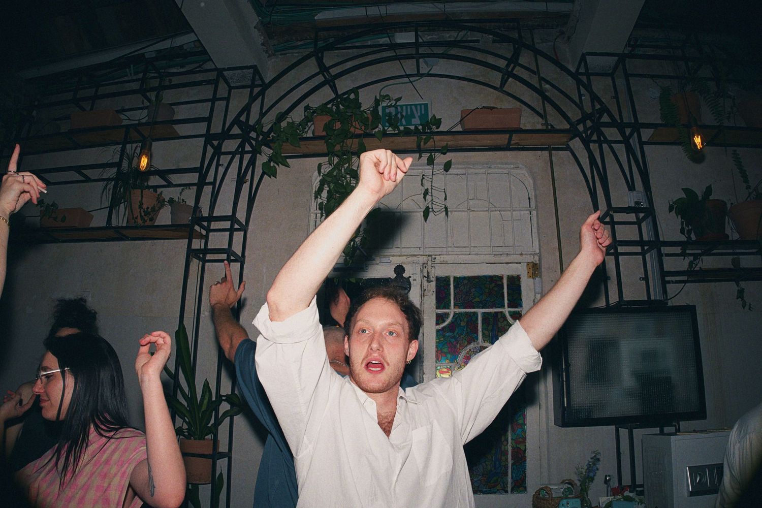 Young person in white shirt dancing with arms raised at an indoor celebration with other guests.