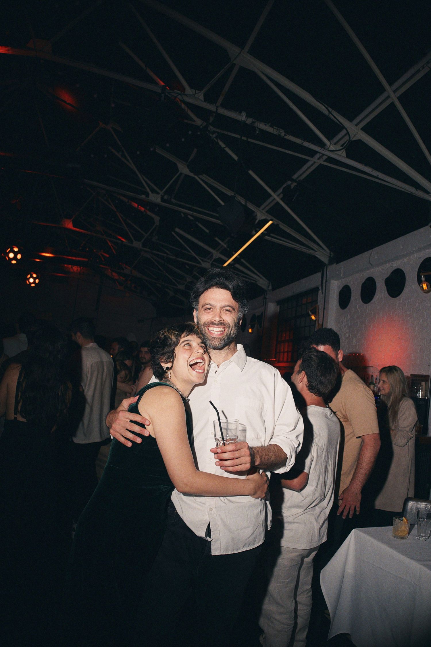 Joyful couple embracing and laughing at lively wedding reception party under tent lighting.