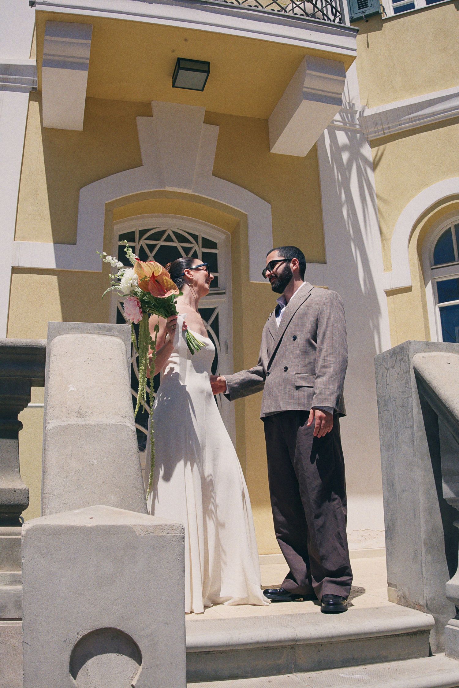 Bride and groom holding hands on steps of elegant yellow building on sunny wedding day.