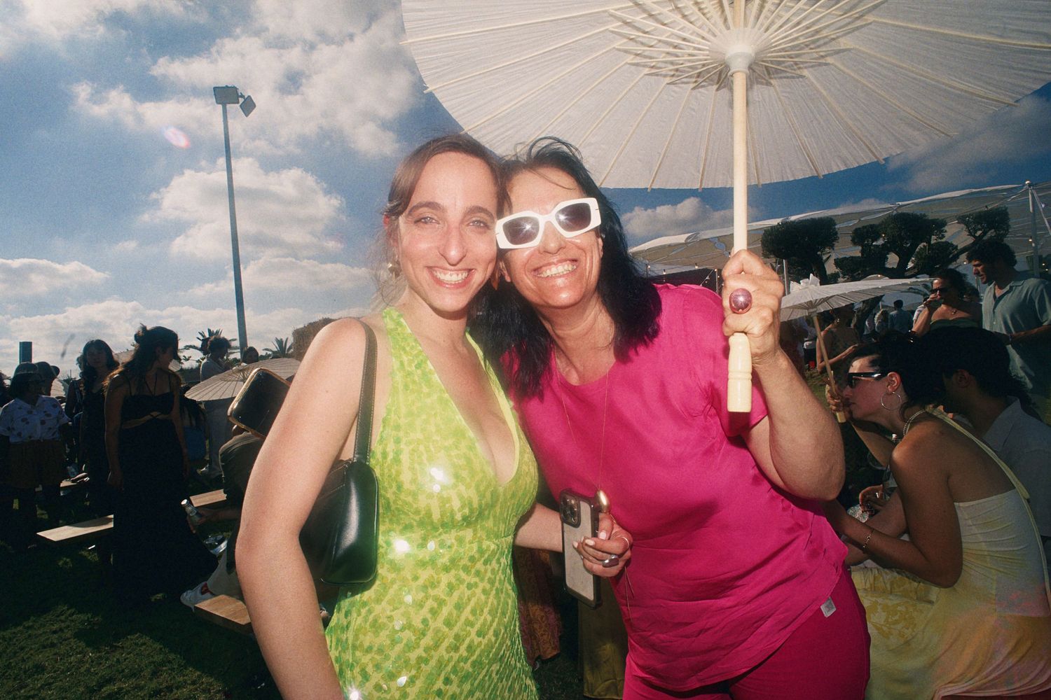Two women smile at outdoor summer event, one in green sequin dress, other in pink holding white parasol.