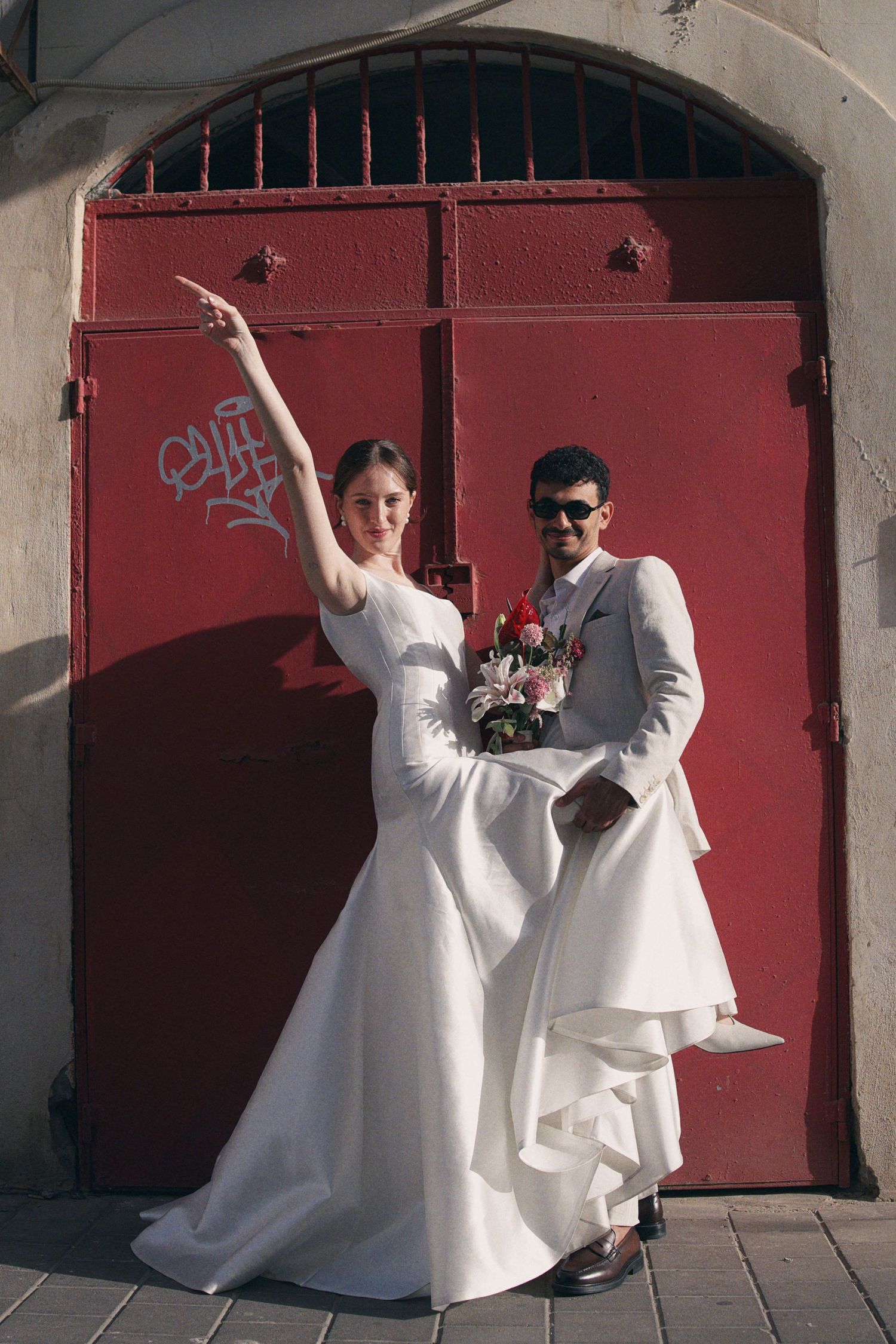 Bride in white mermaid gown and groom in white suit pose playfully before a red door with graffiti.
