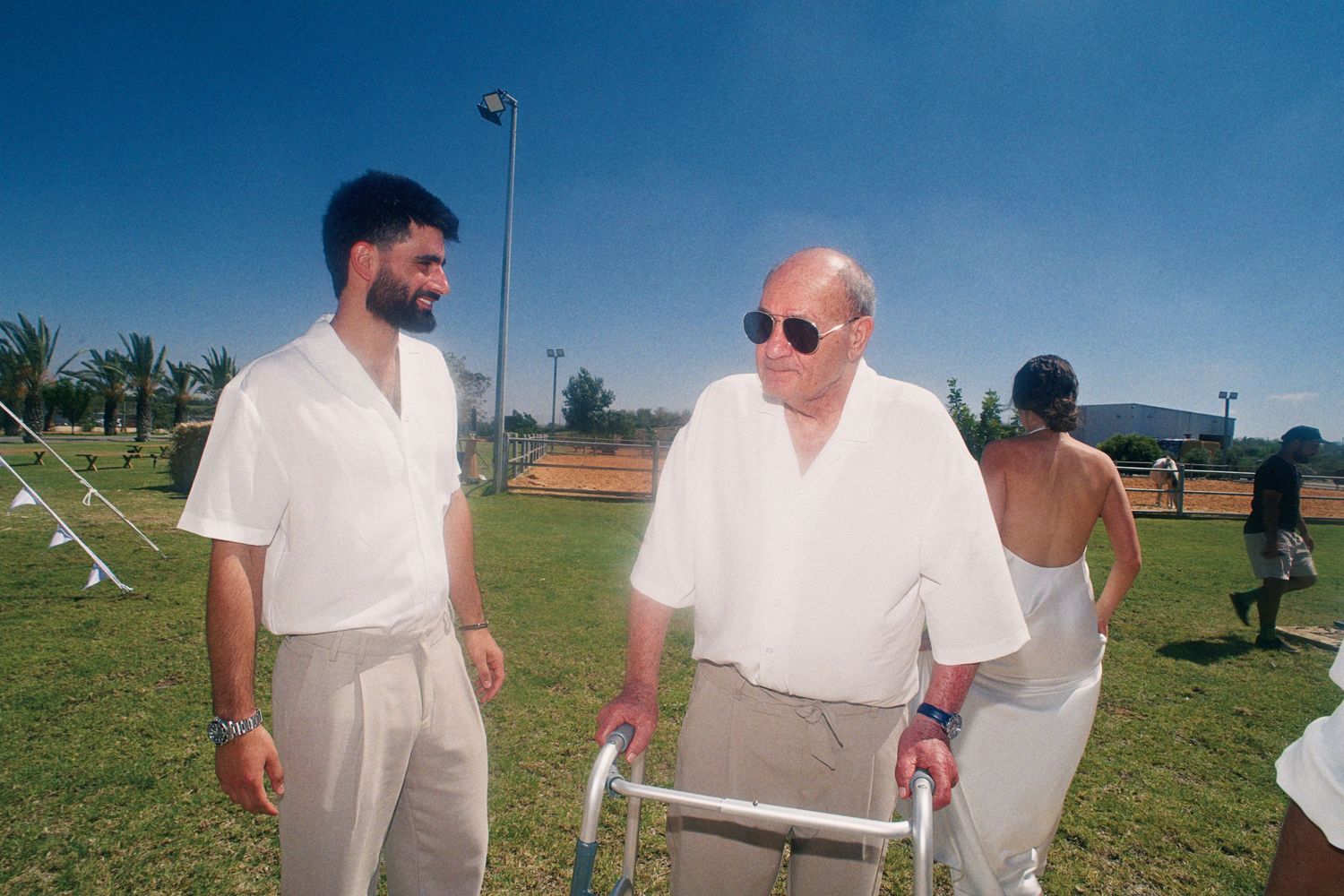 Two men in white shirts chat outdoors on a sunny day, one using a walker, with palm trees in the background.