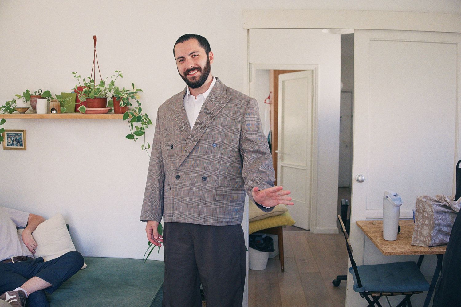 Smiling bearded man in oversized gray plaid blazer gesturing in a modern apartment living room.