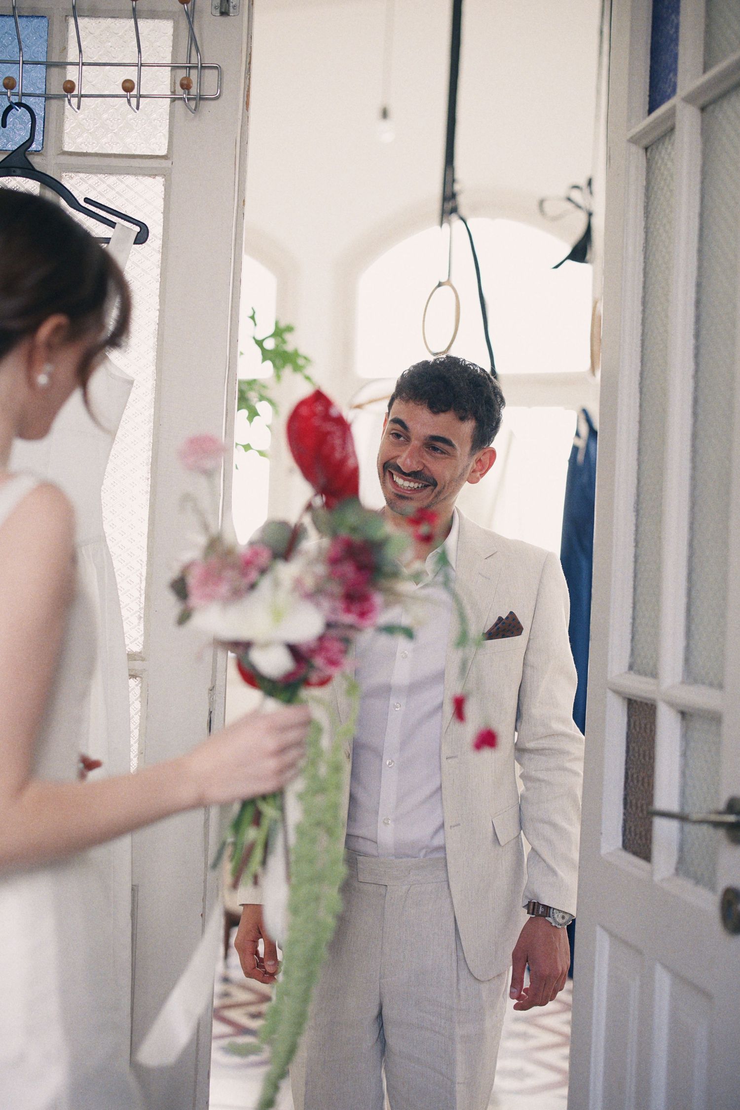 Smiling groom in beige suit reacts to bride holding colorful wedding bouquet in bright doorway.