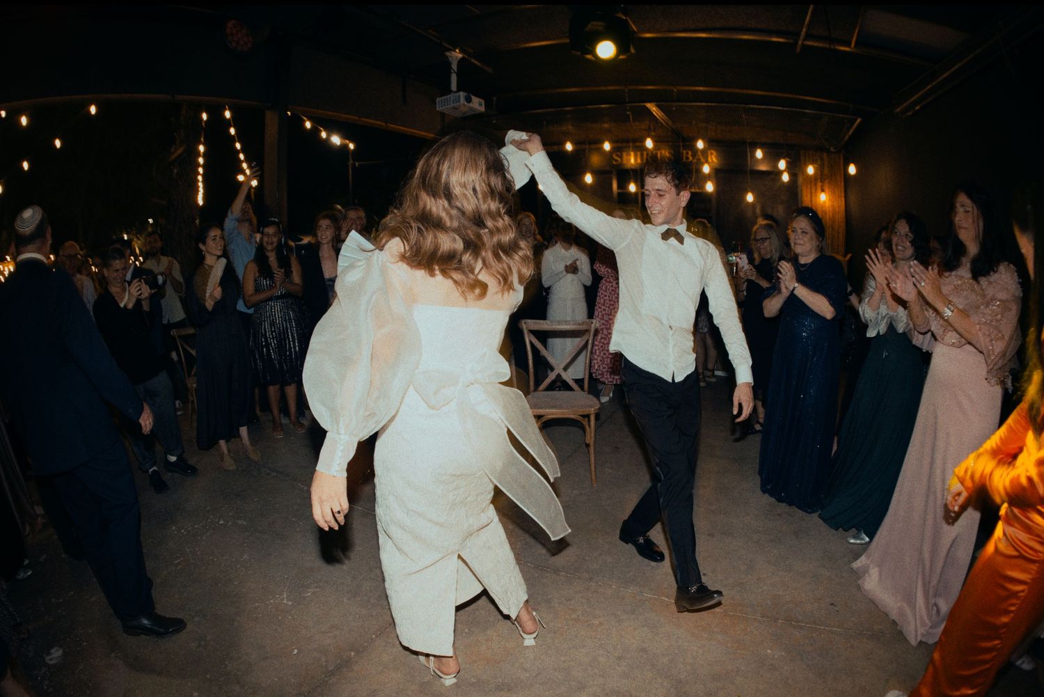 Dancers in formal attire twirl and move energetically on a dimly lit dance floor at a celebration.