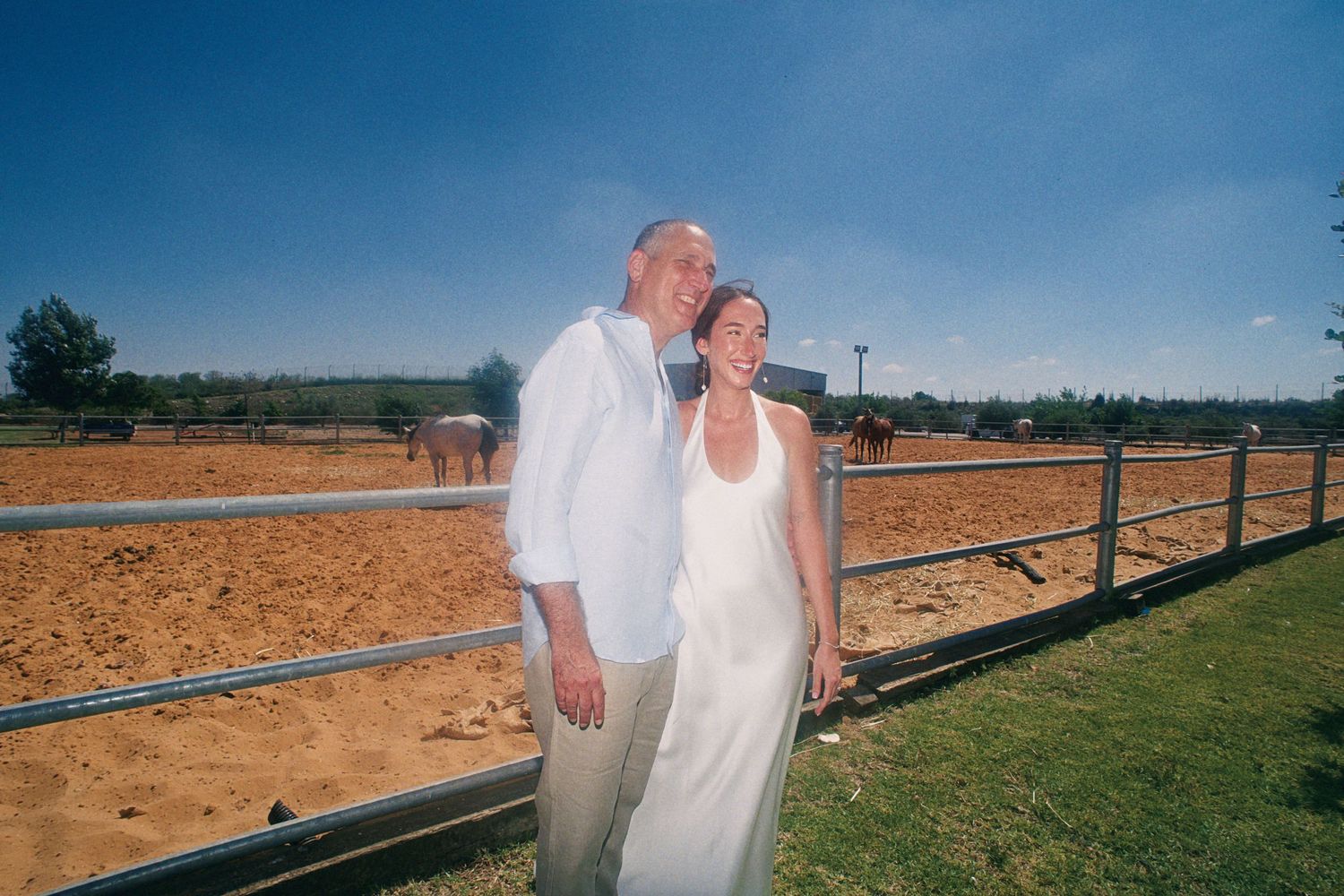 A couple in wedding attire smiling together at a sunny horse ranch with a sandy paddock and metal fence.