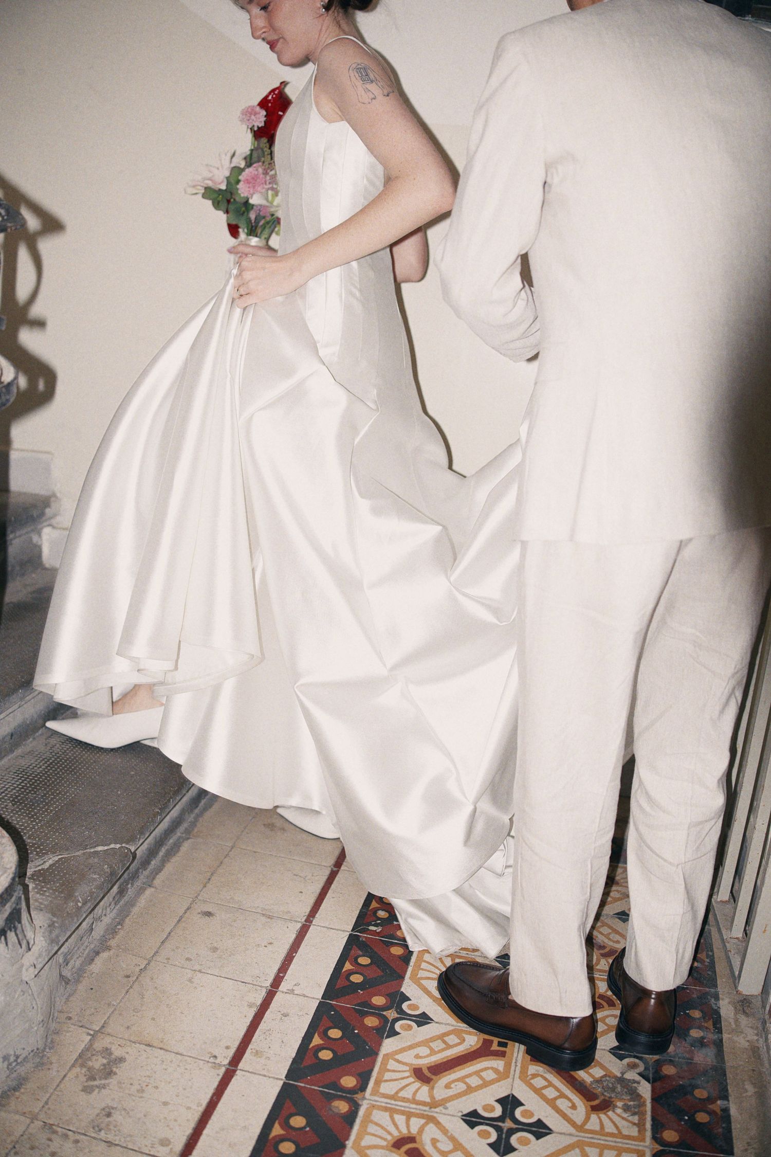 Bride in white ball gown holding red bouquet walks up stairs assisted by groom in beige suit.