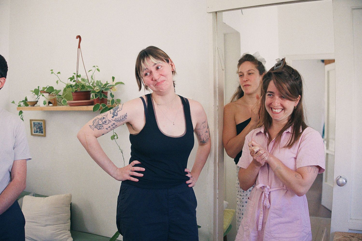 Three women socializing indoors, one with tattoos posing confidently, another laughing in a pink dress.
