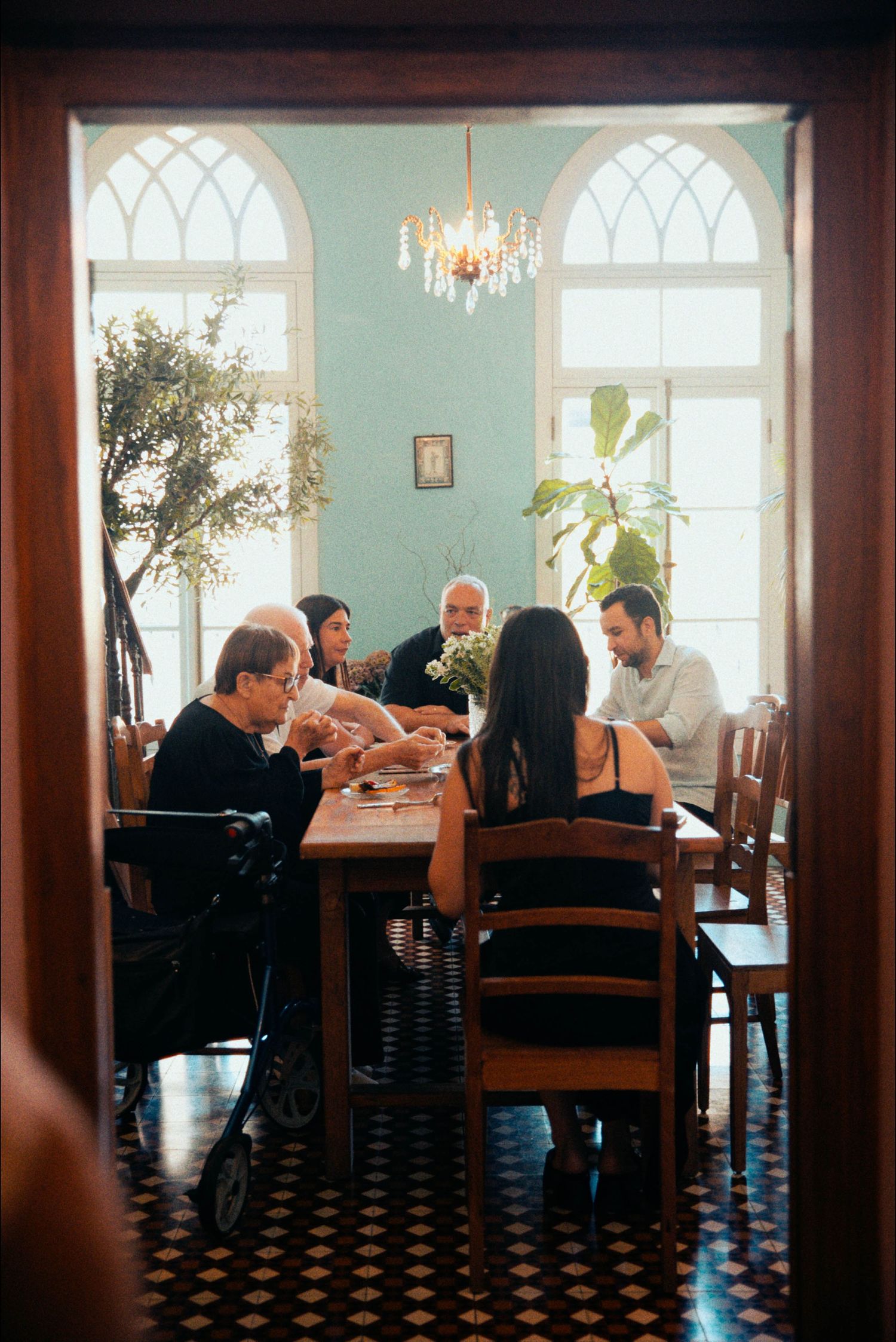 A group gathers around a dining table under elegant arched windows and a crystal chandelier.