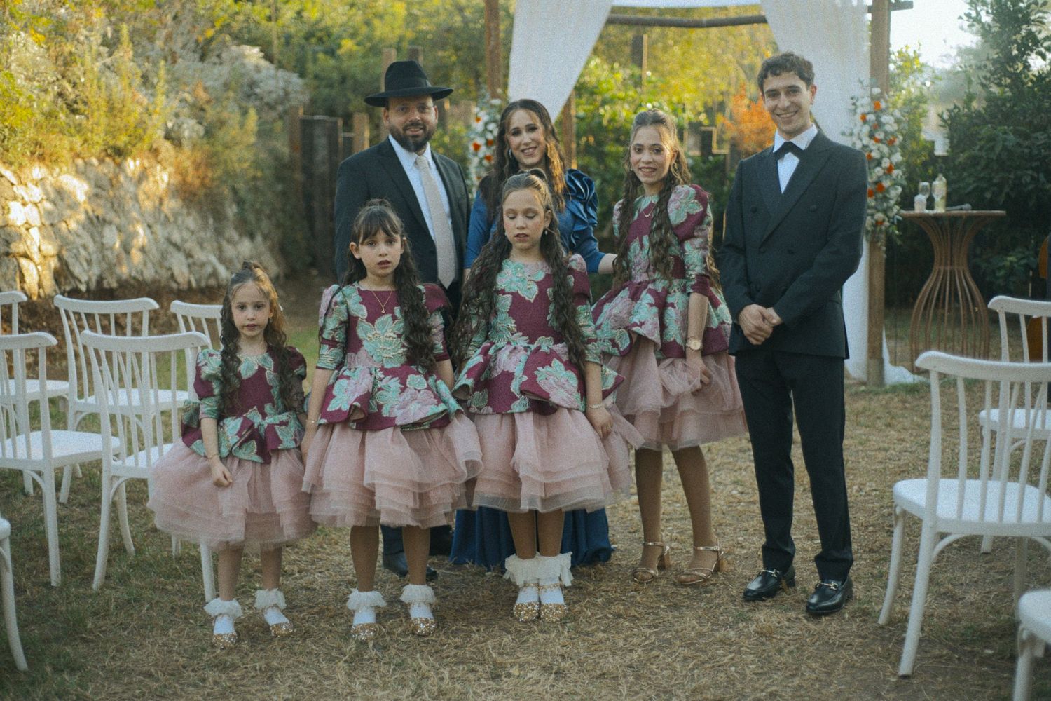 Wedding guests and children in formal attire gather under white outdoor ceremony setup.