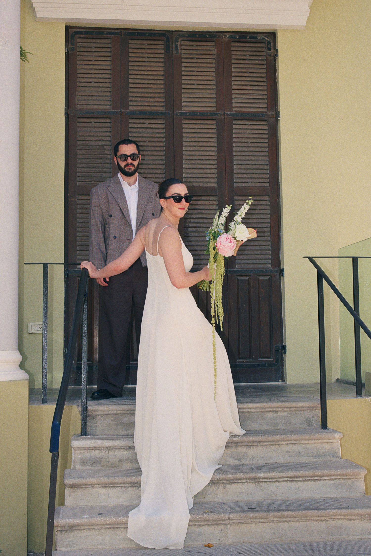 Bride in elegant white chiffon gown holds bouquet on steps with groom in gray suit, both wearing sunglasses outdoors.