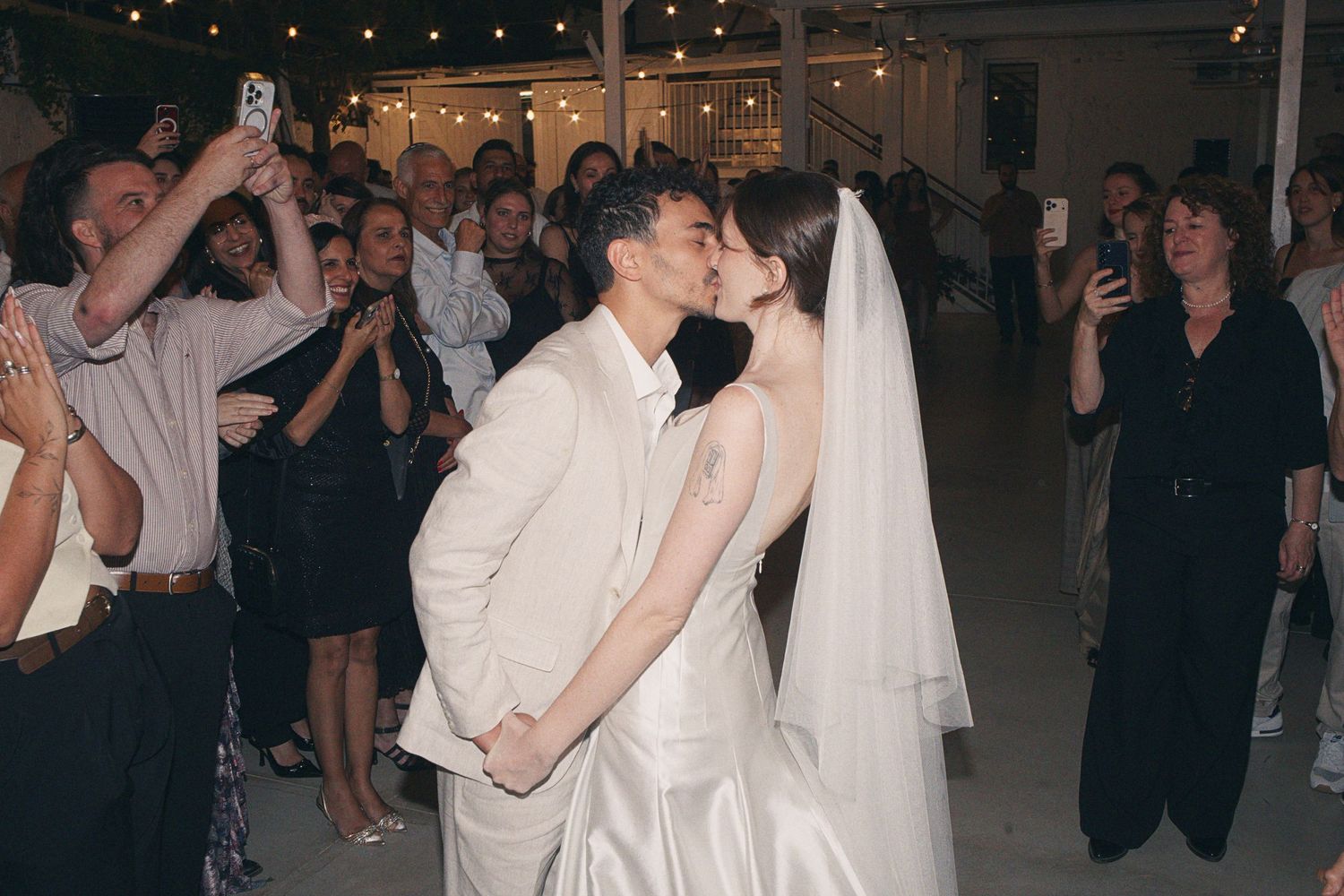 Bride and groom share a first kiss at their wedding reception surrounded by cheering guests taking photos.