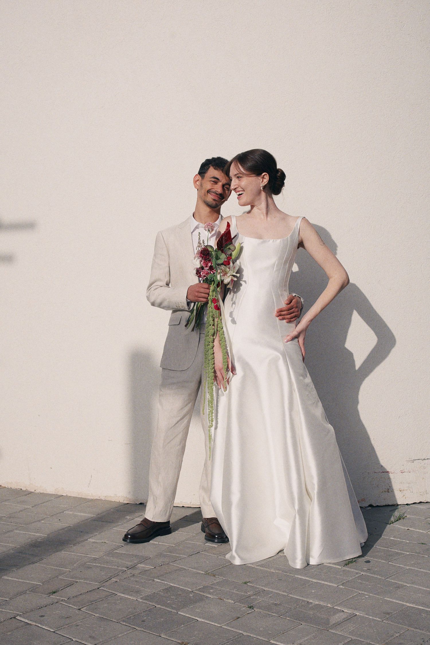 Bride in white mermaid gown and groom in beige suit posing with red bouquet against white wall.