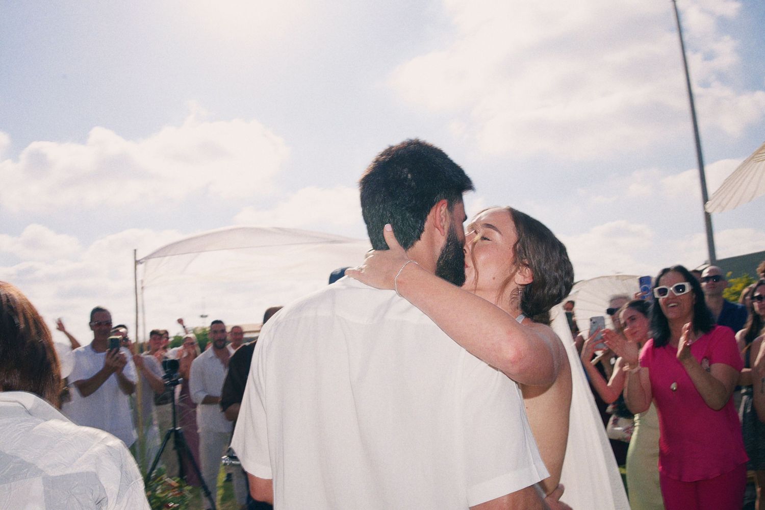 A couple shares their first kiss as newlyweds outdoors, surrounded by cheering guests under a bright cloudy sky.