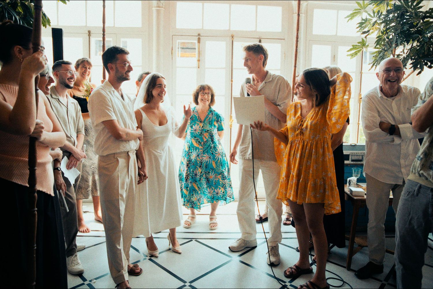 Wedding guests mingling and dancing in a bright indoor venue with large windows.