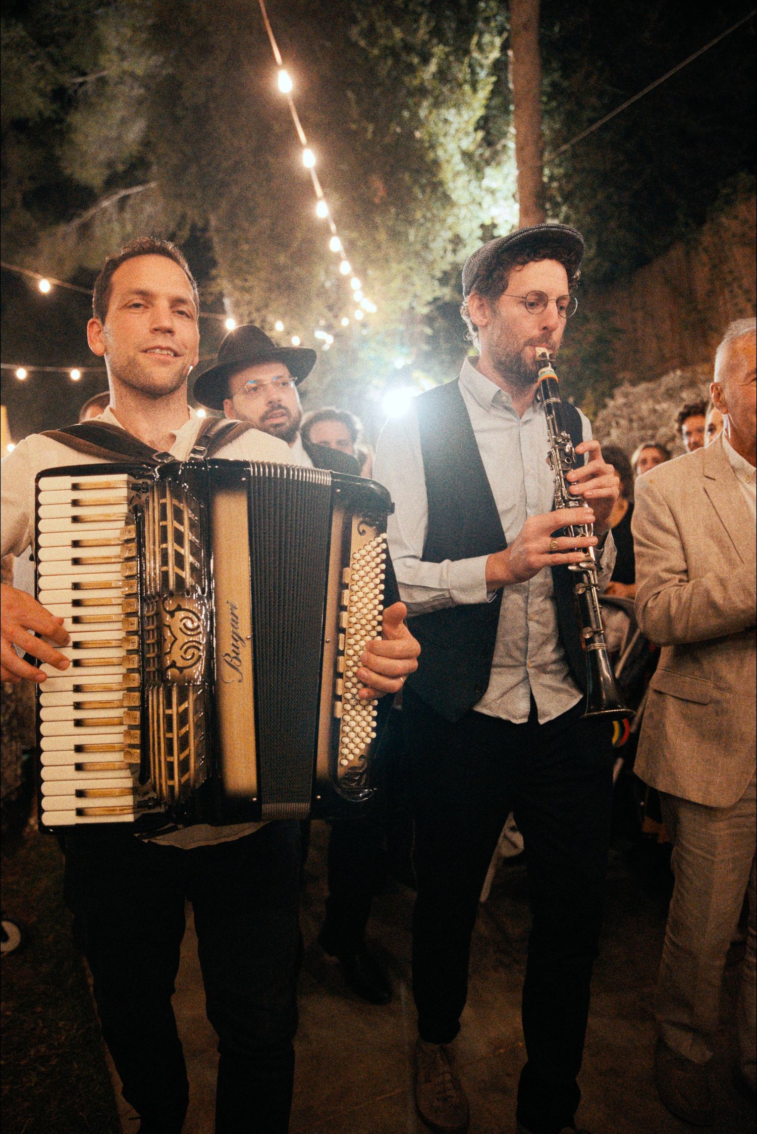 Musicians play accordions at a nighttime celebration with string lights overhead.
