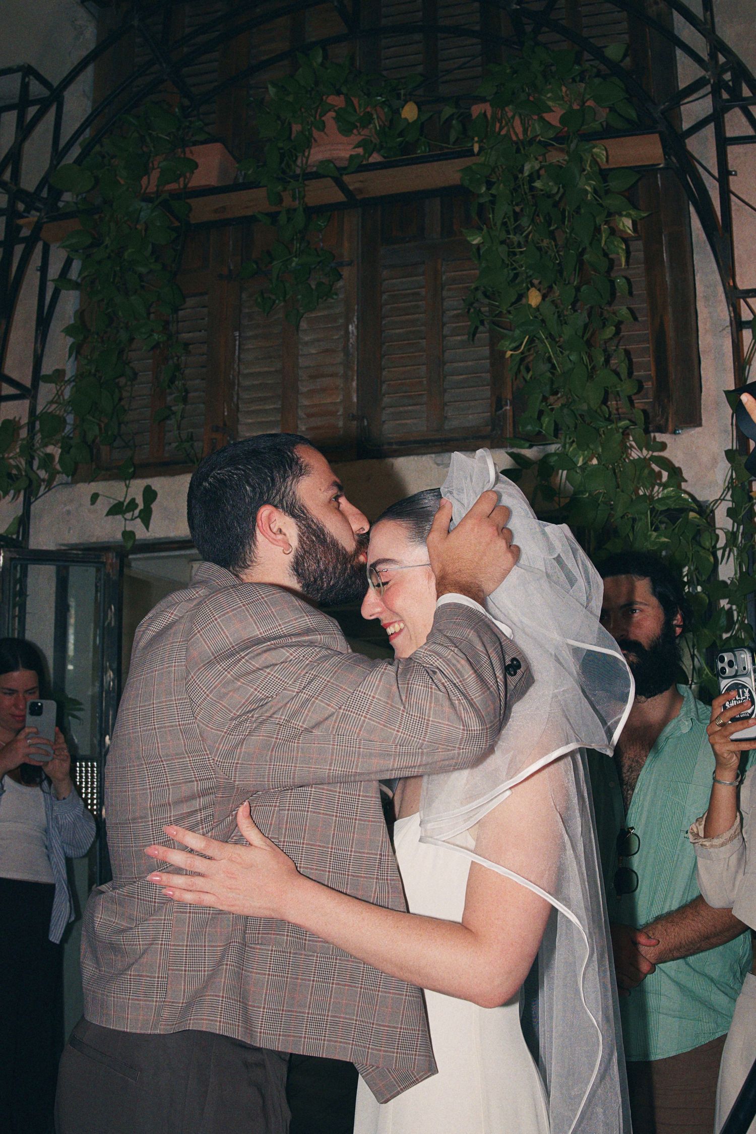 Newlyweds share first kiss under green floral arch surrounded by wedding guests.