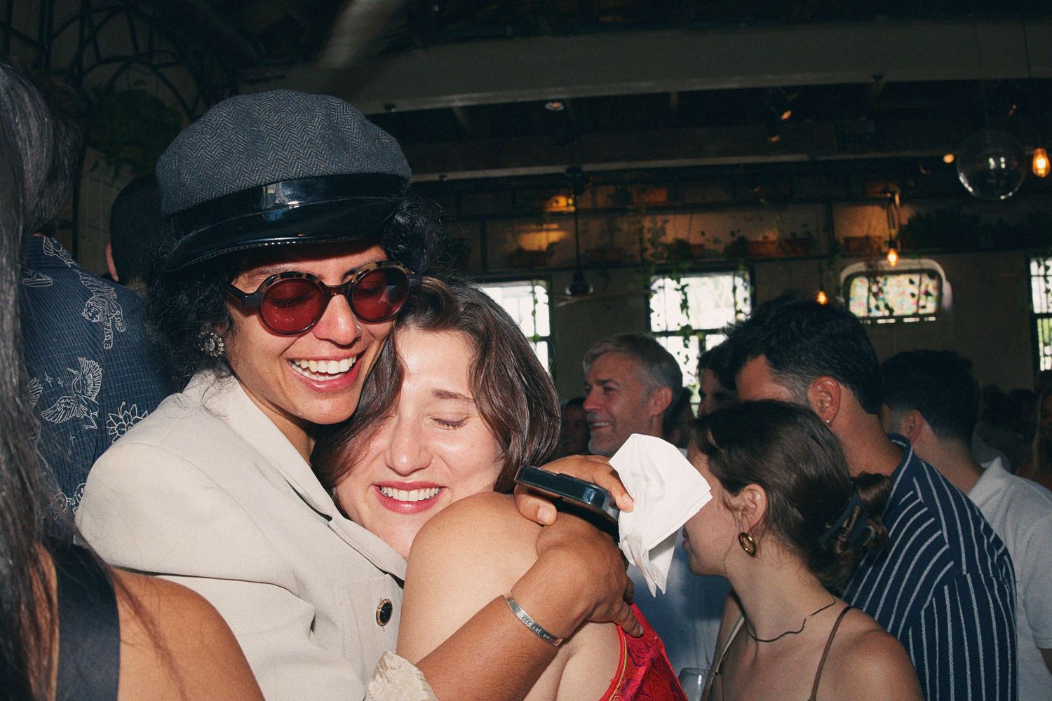 Two friends hugging and smiling at indoor party, one wearing hat and sunglasses.