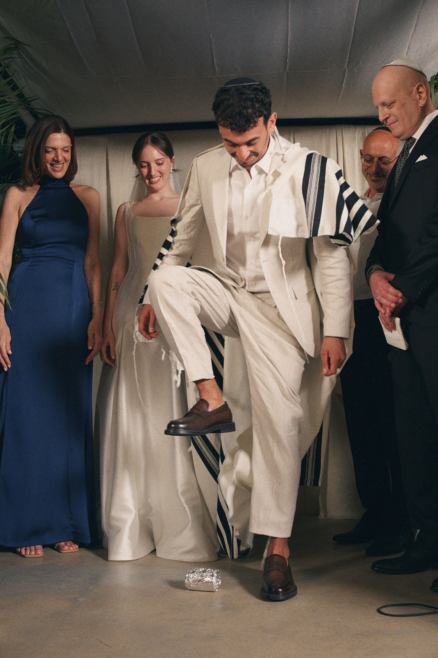 Groom in white suit performs Jewish glass breaking ceremony surrounded by smiling wedding guests.
