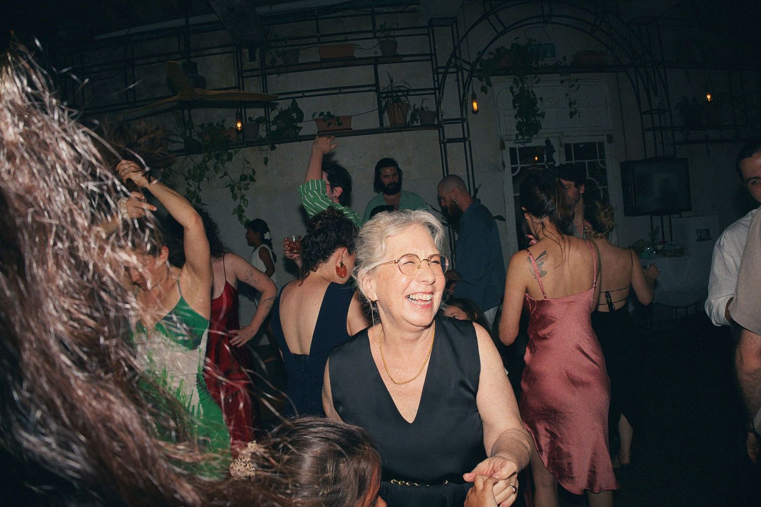 Elderly woman smiling and dancing joyfully at a lively indoor party with guests in formal attire.