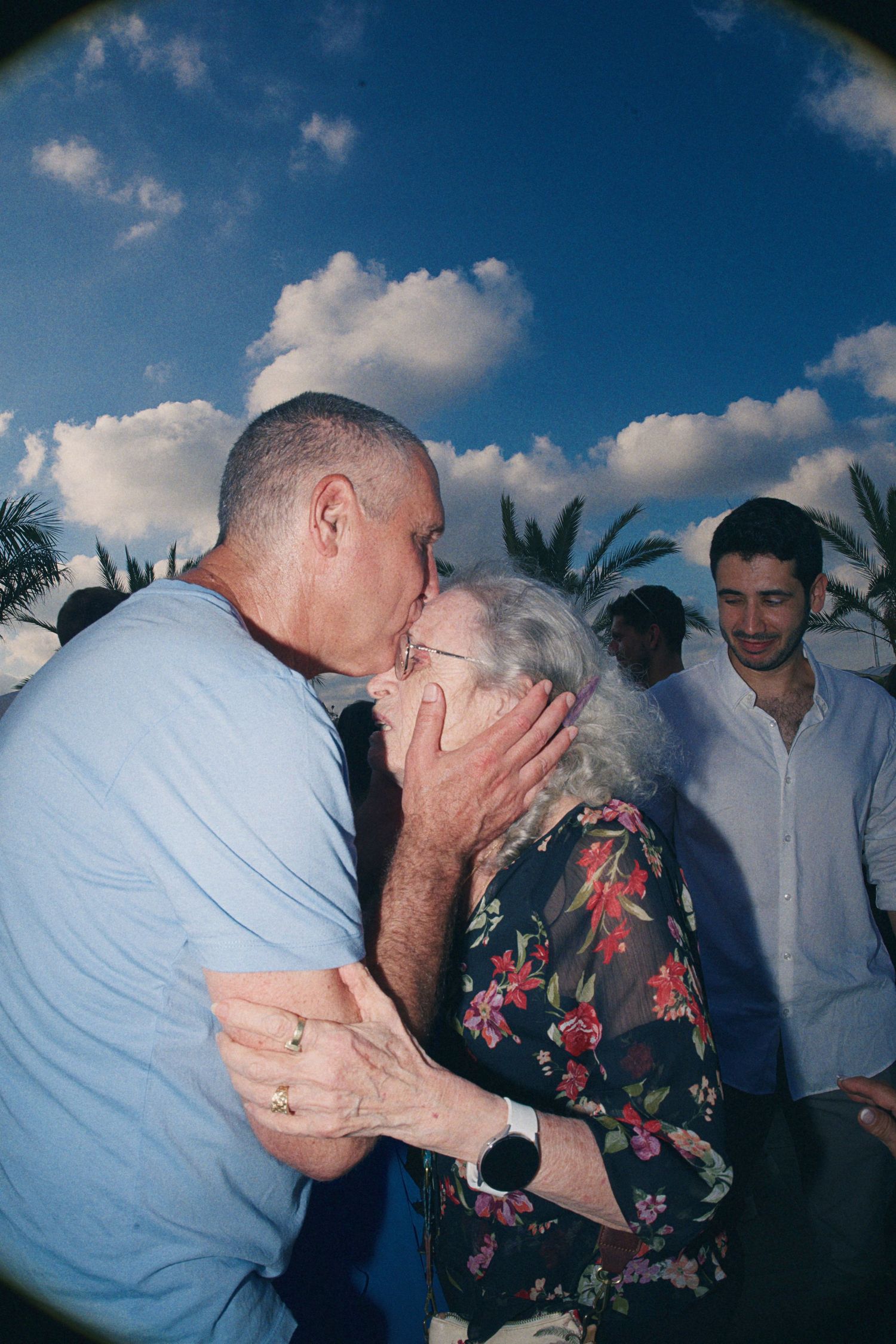 Elderly couple sharing a tender kiss at an outdoor event with palm trees and blue sky.