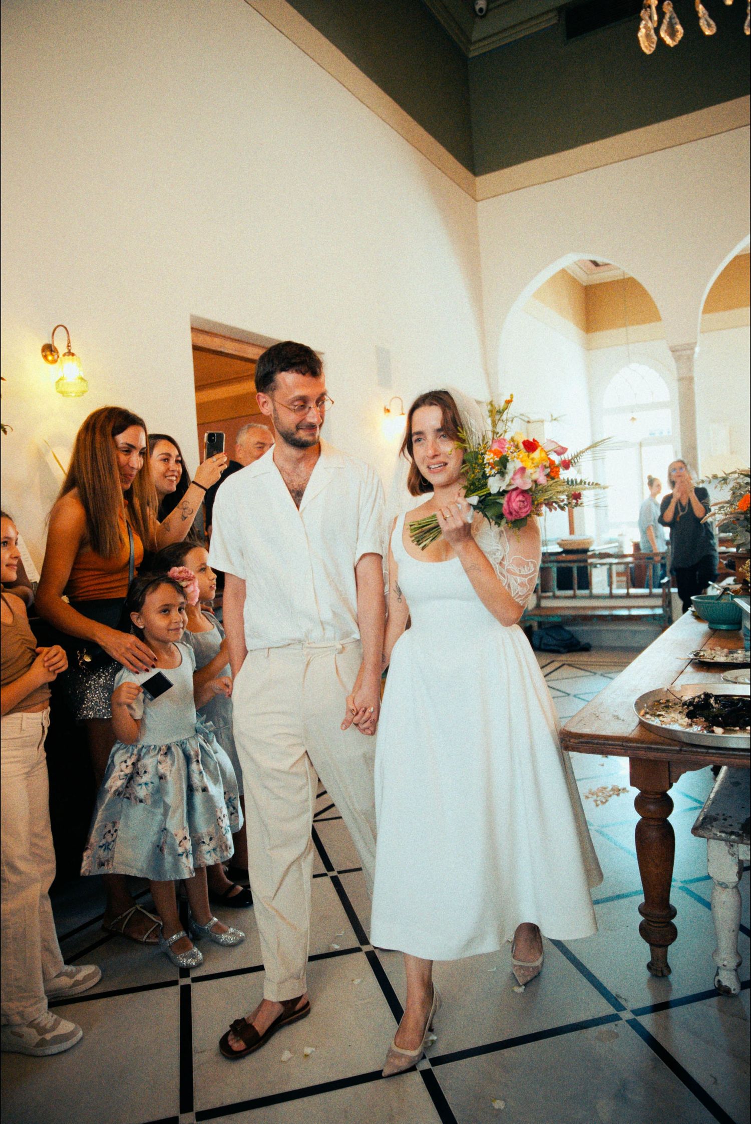 Newlyweds in white outfits stand by a dessert table at their reception.