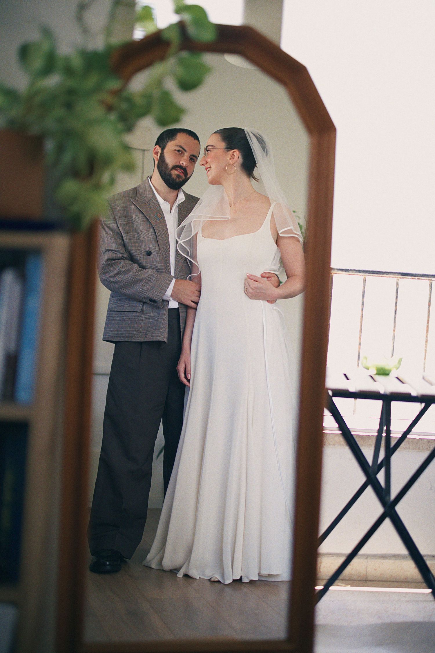 Bride and groom share an intimate moment reflected in a wooden mirror, she wearing a flowing white gown with veil.