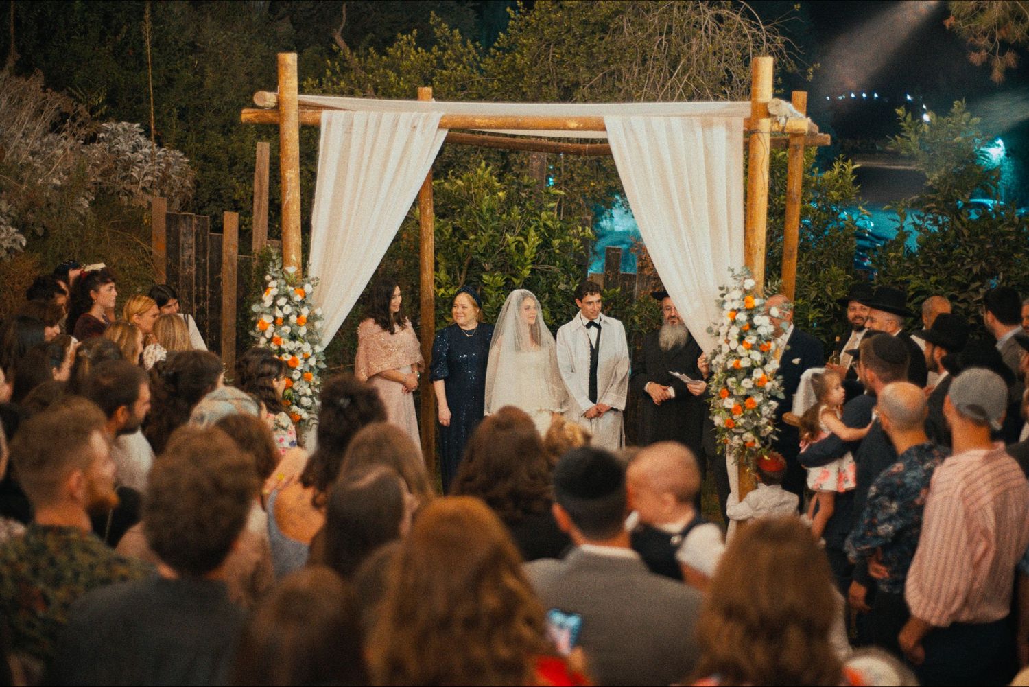 Outdoor evening wedding ceremony with guests gathered around wooden arch decorated with white flowers.