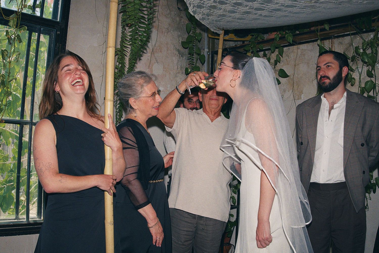 Bride in veil drinking from cup during Jewish wedding ceremony under chuppah with guests laughing.