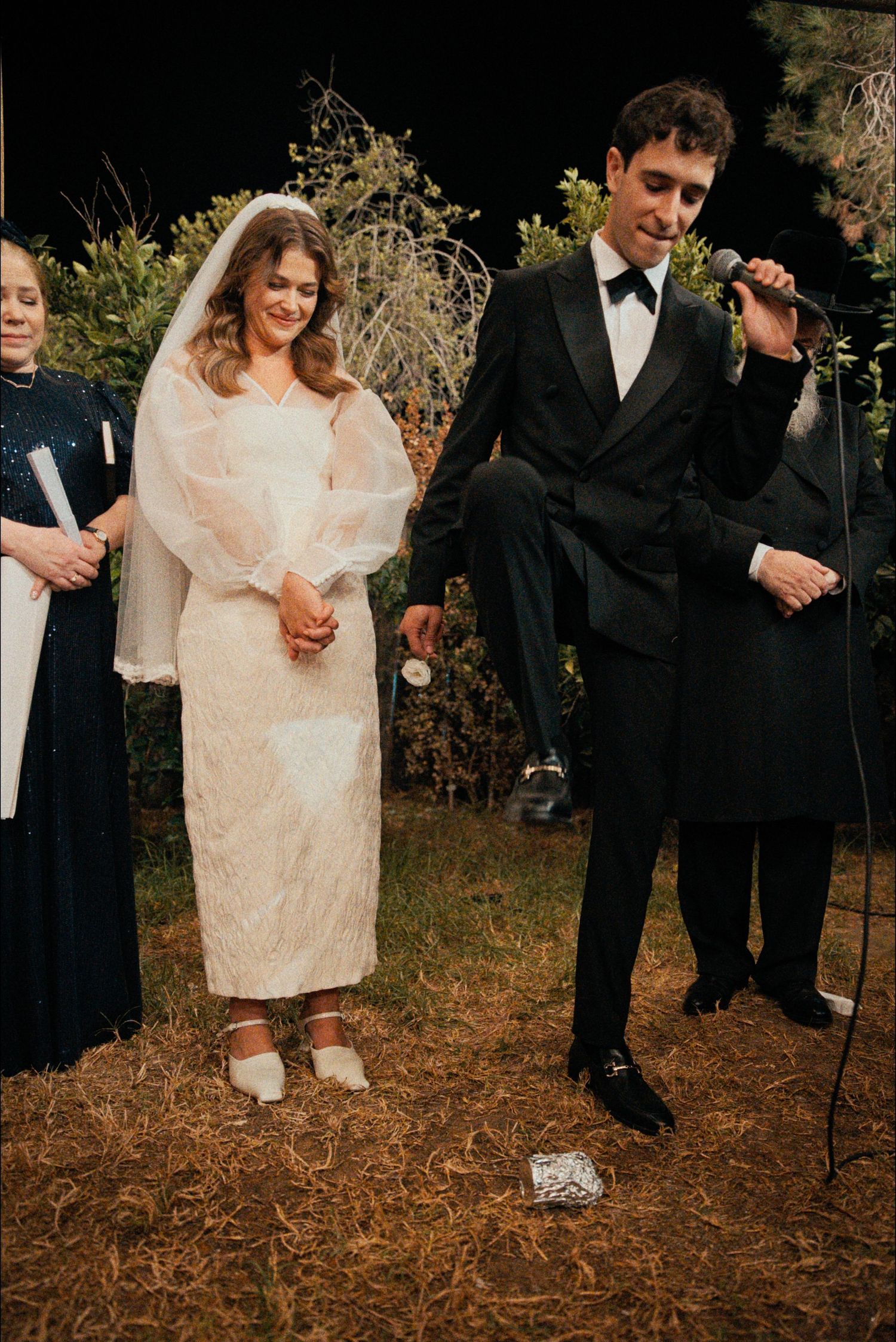 Wedding celebration participants stand together on autumn leaves in formal attire.
