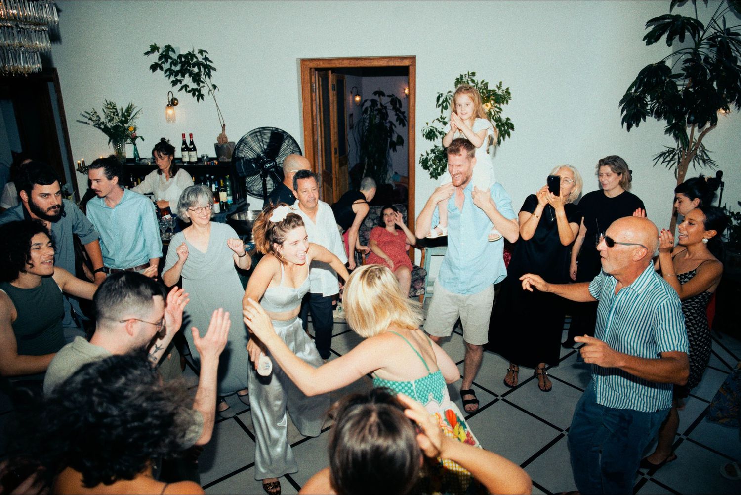 Party guests dance and socialize in a bright indoor space with plants and decorations.
