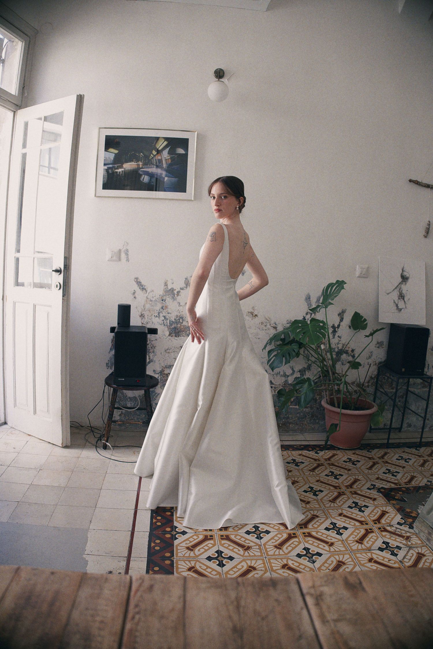 Elegant bride in a white backless satin wedding gown standing in a vintage room with patterned tile floors.