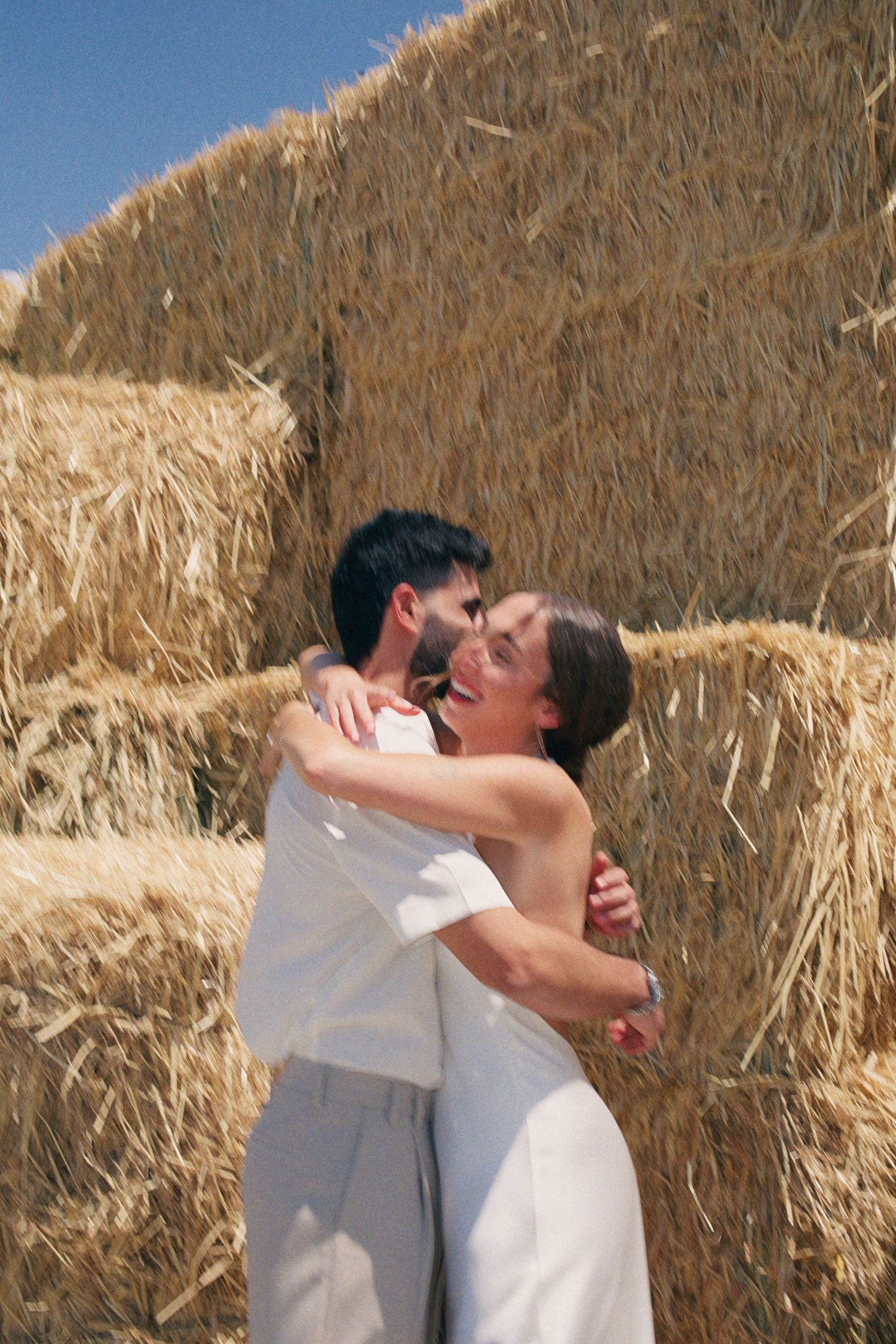 Couple embracing and laughing in front of golden hay bales on a sunny day.