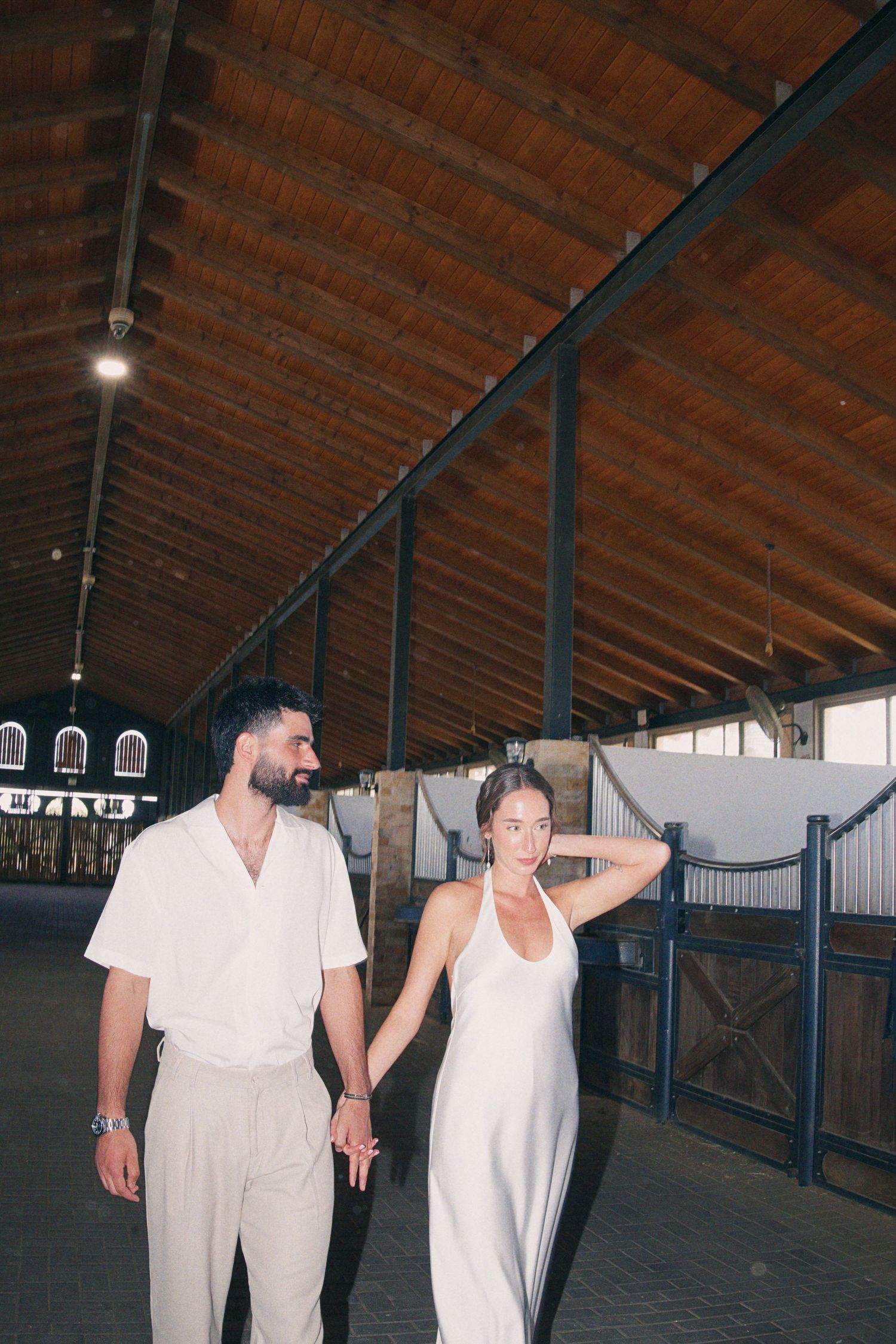 Bride and groom in white outfits hold hands walking through a rustic wooden stable venue.