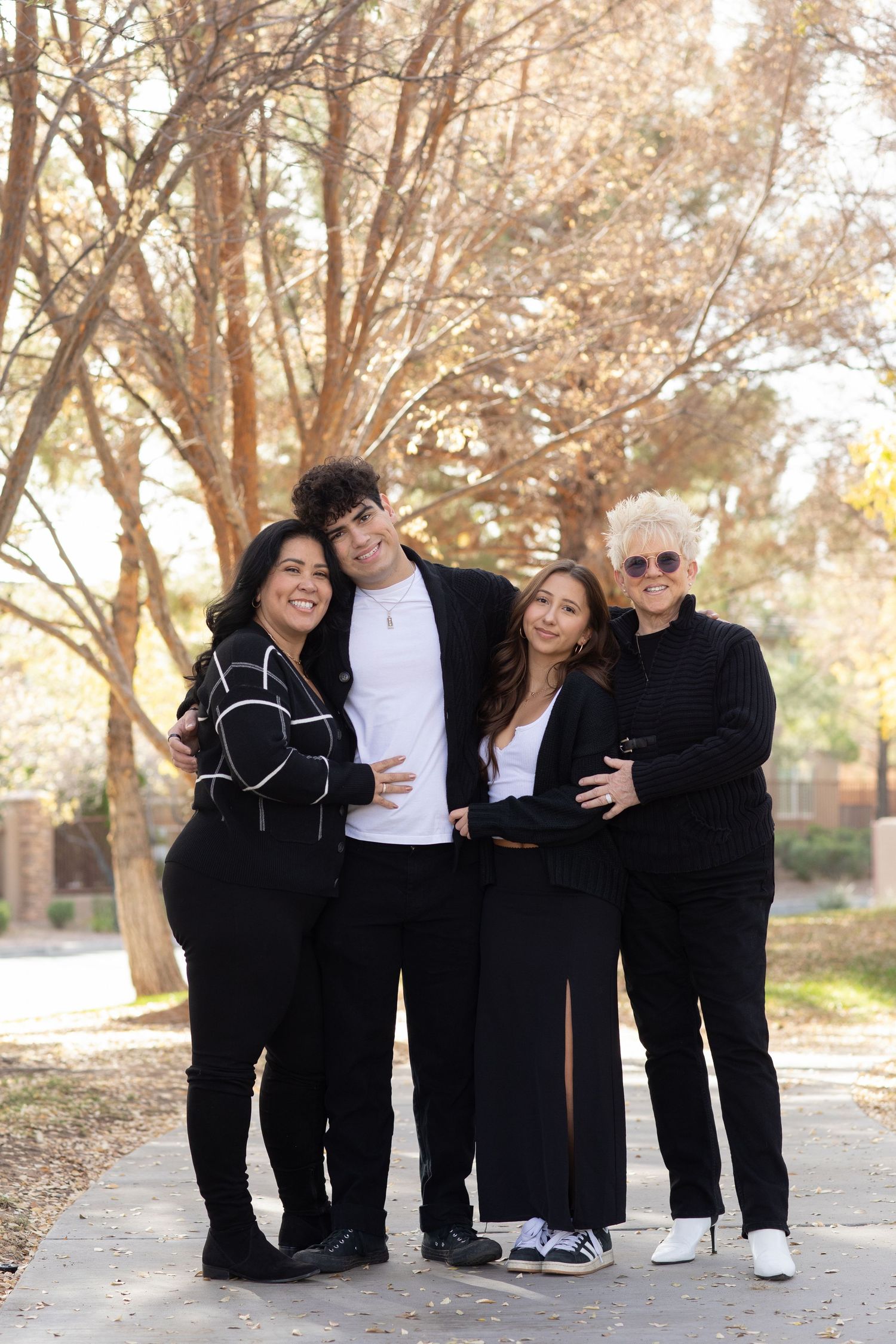 A group of four people in black outfits pose together outdoors with autumn trees in the background.