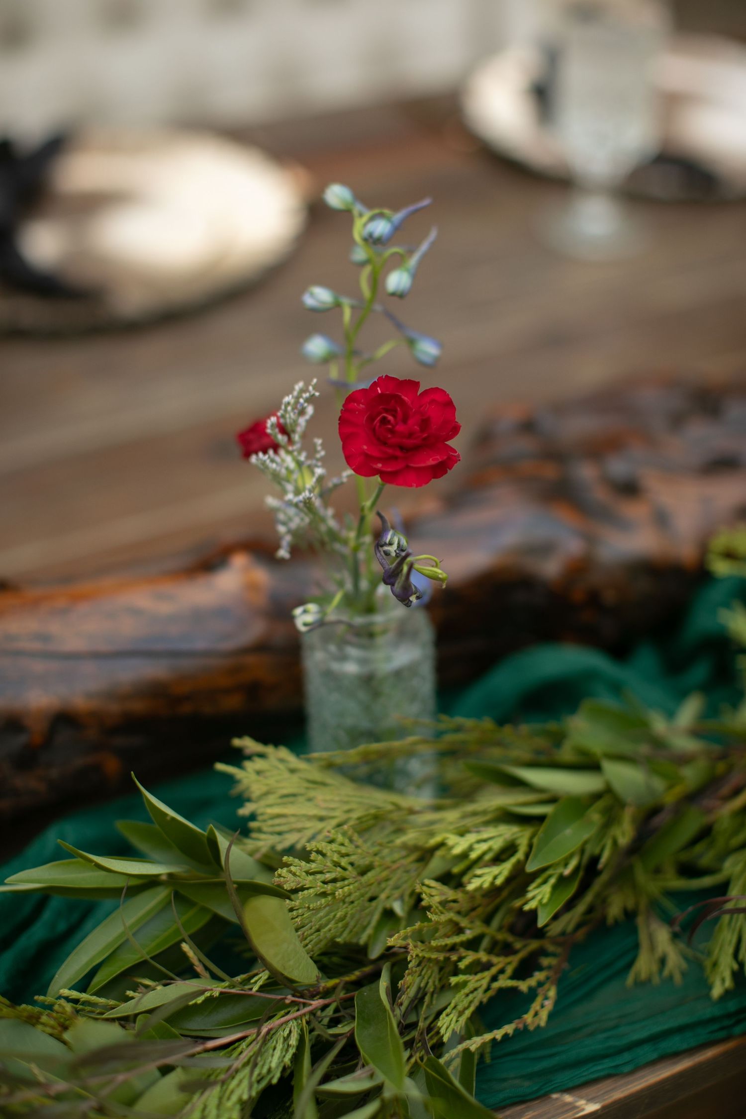 Wedding table decor featuring rustic greenery and a single red rose in a glass vase.