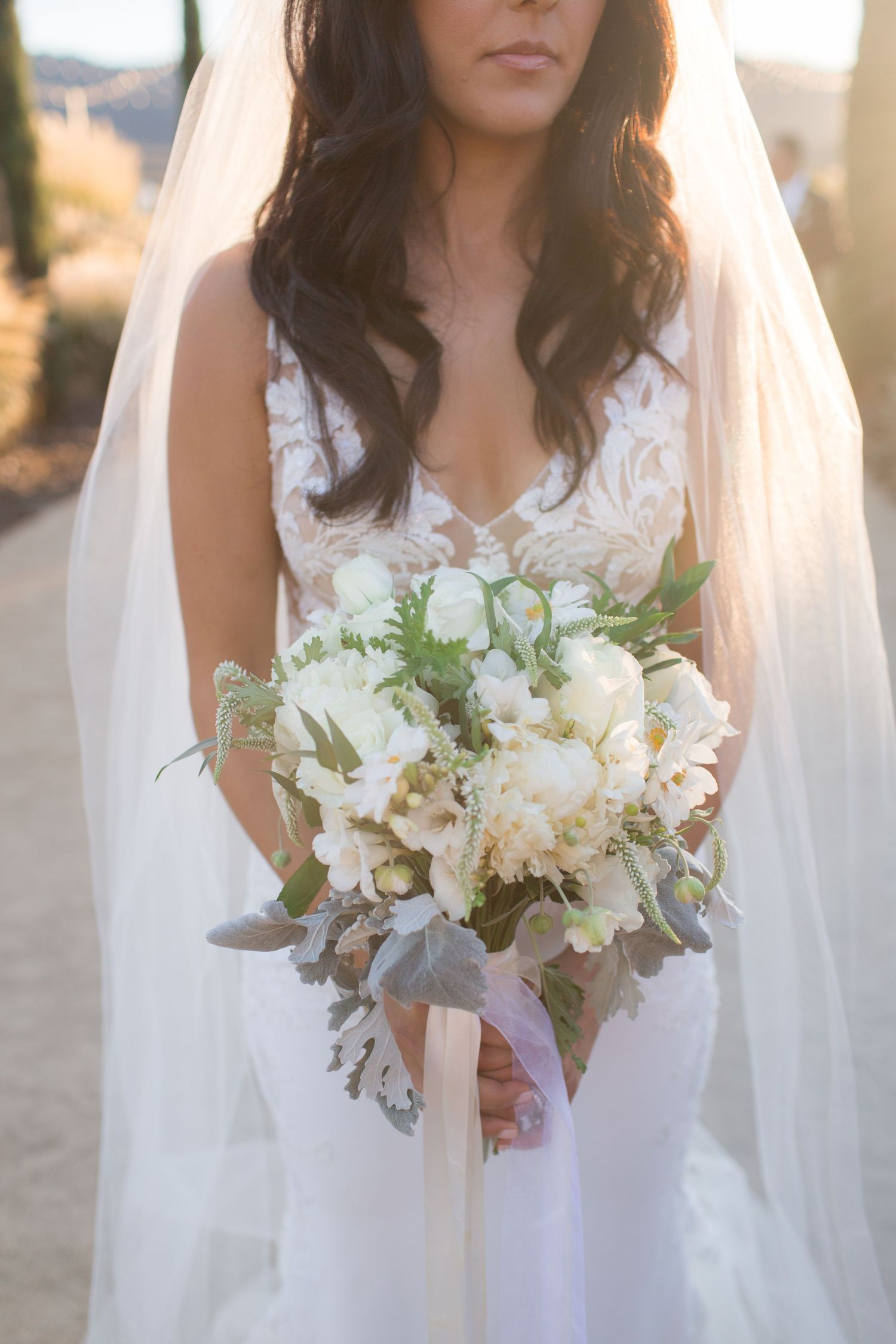A bride in a lace wedding dress holds an elegant white and green bouquet against a warm sunset background.