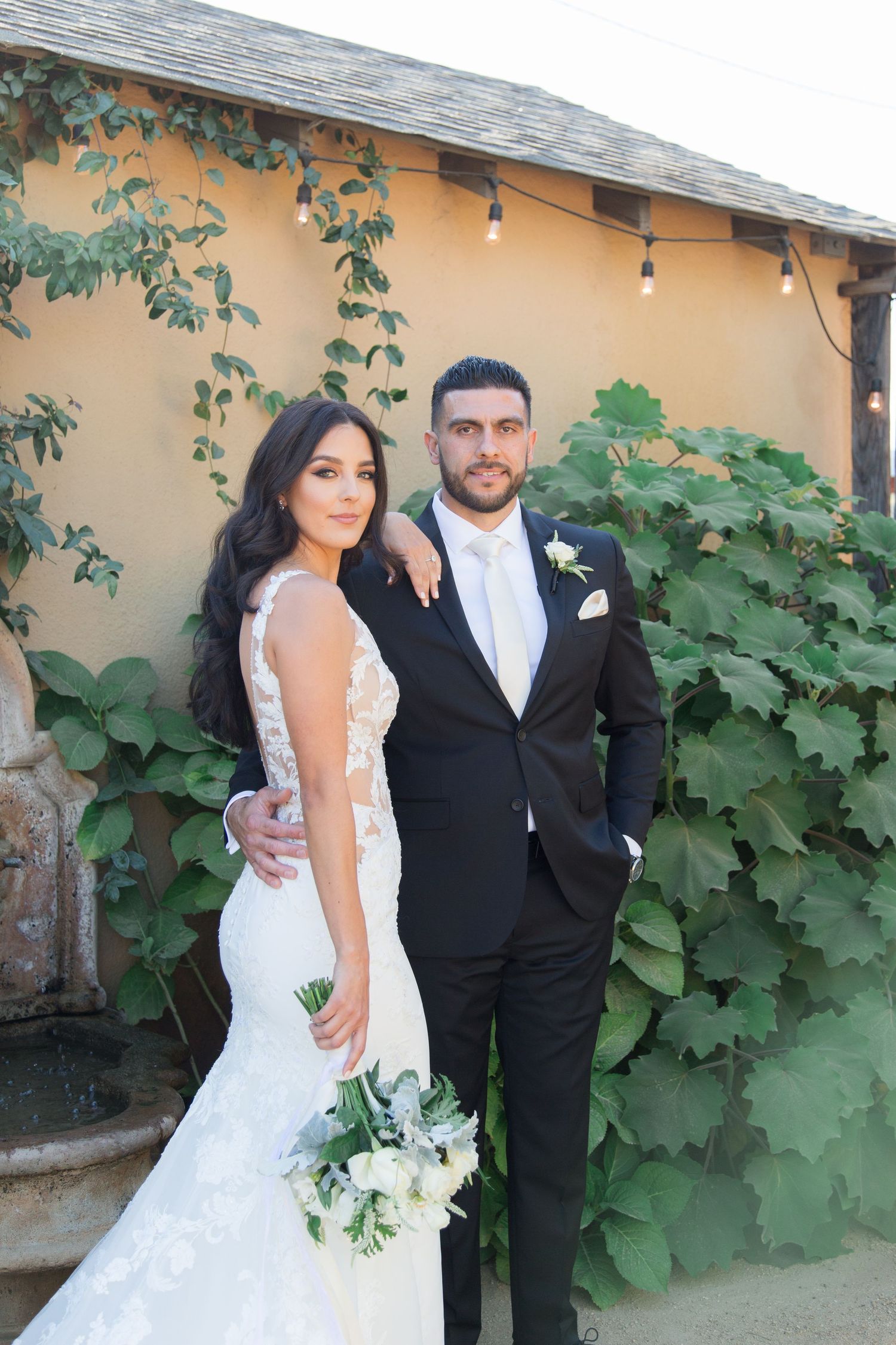 A bride in a fitted lace gown and groom in black tuxedo pose against a yellow stucco wall with climbing vines.