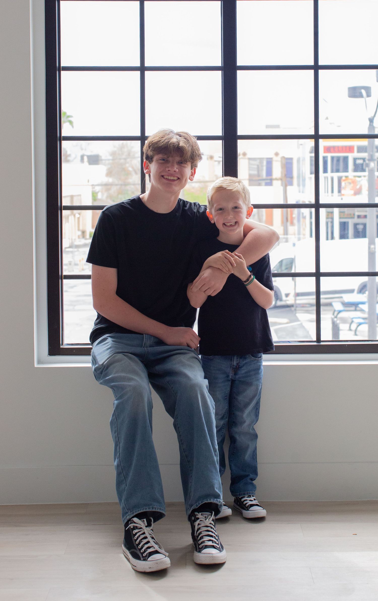 Two people wearing casual clothes and sneakers sit by a window in a bright interior space.