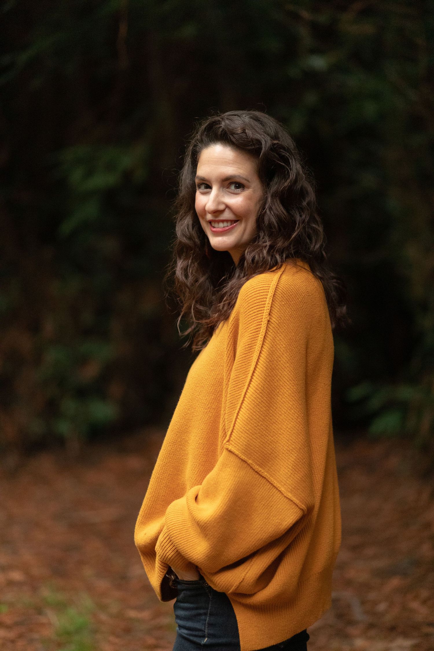Person in mustard yellow sweater poses playfully against dark forest background.